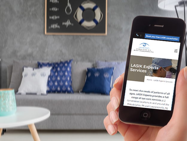 A right hand holding a phone with LASIK Experts website on the screen and a living room backdrop with couch and pillows. Both the phone and backdrop are slightly out of focus.