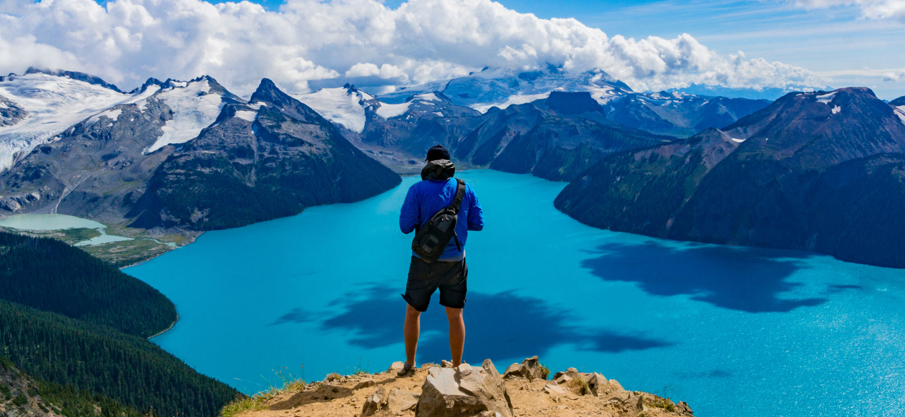 A man wearing a backpack stands on a mountaintop overlooking a panorama of water and other mountains.